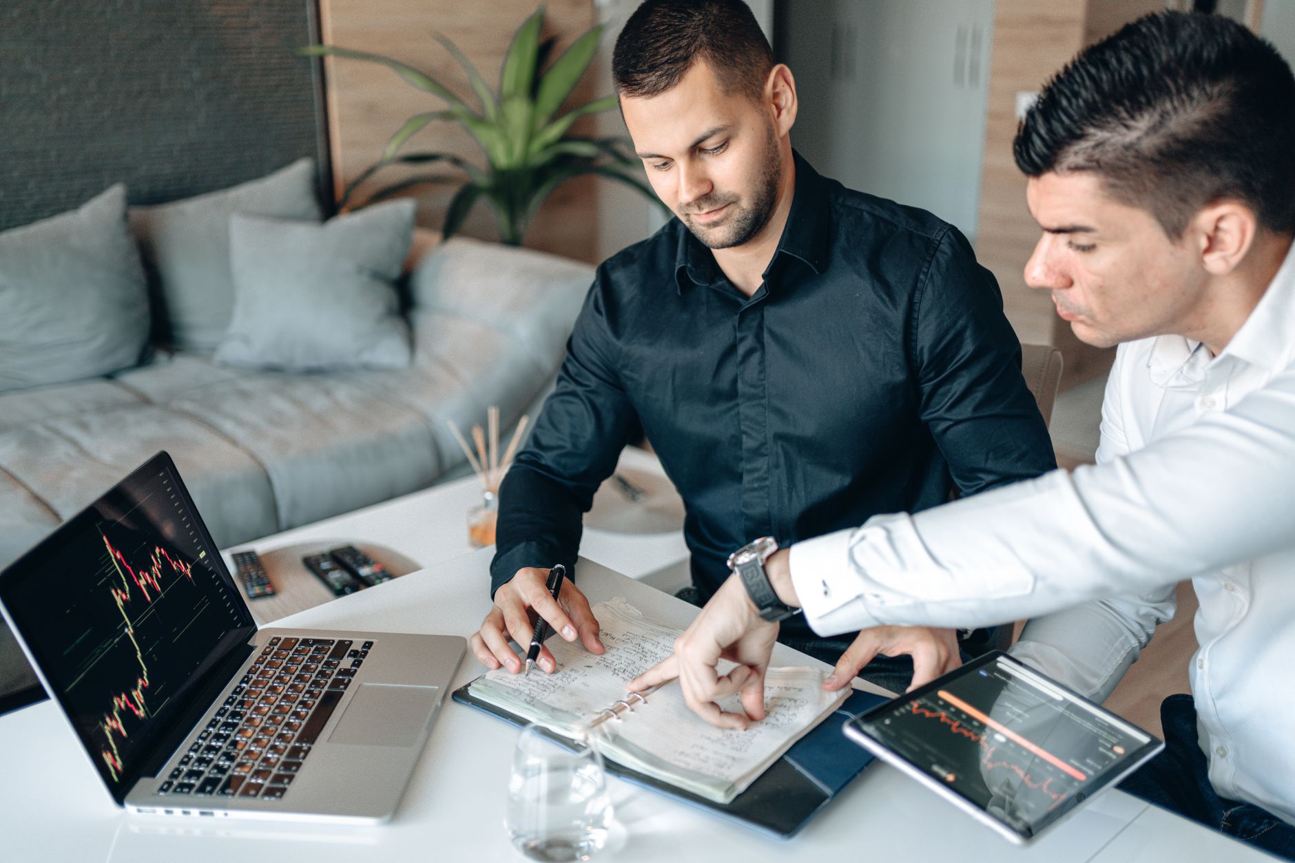 Ilustrasi: Two businessmen collaborating on stock market analysis with charts on laptops and tablets in a modern office.