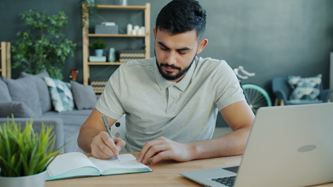 Ilustrasi: Man writing in notebook at desk with laptop.