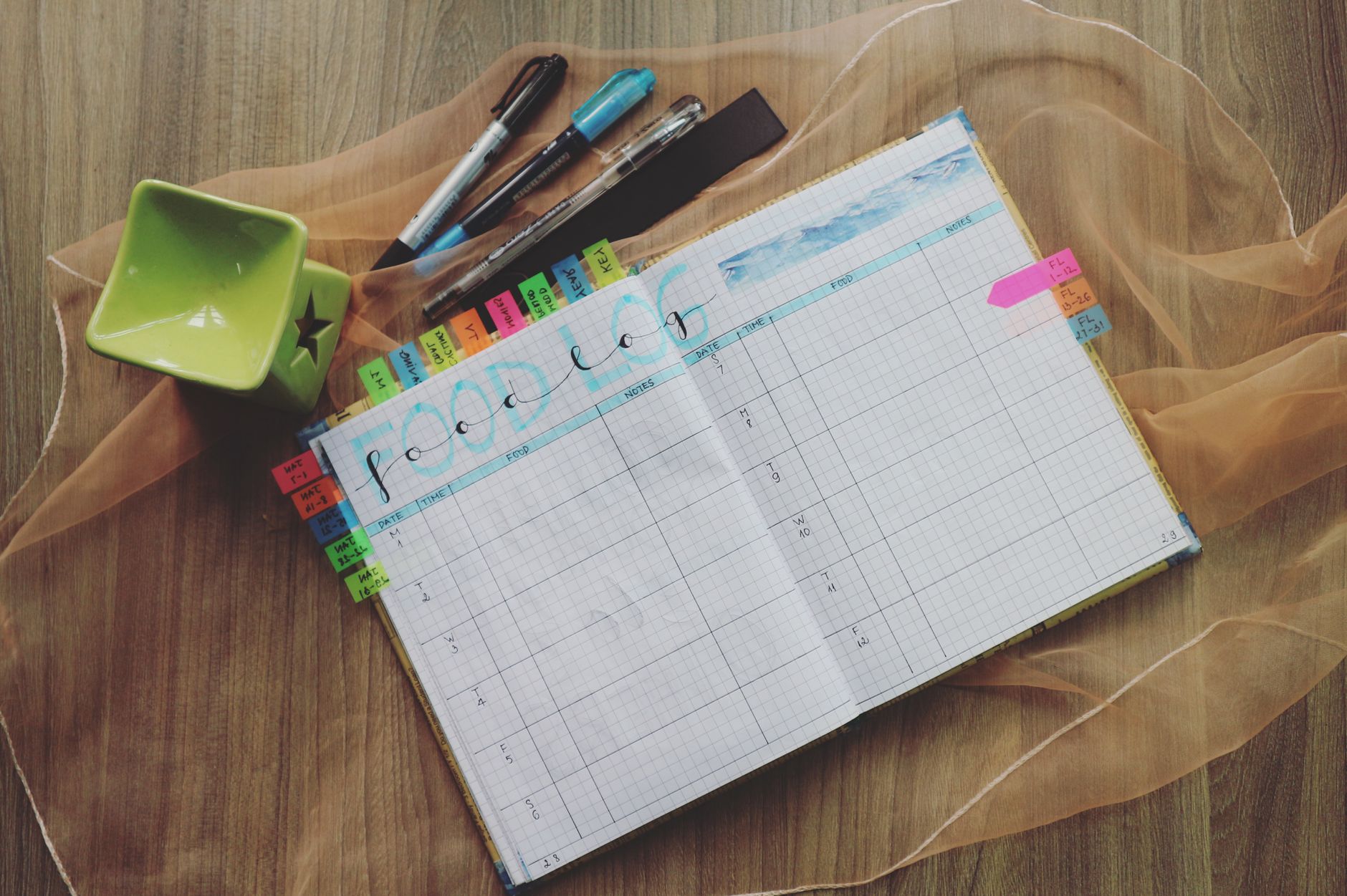 Ilustrasi: Open food log planner with colorful tabs and pens on a wooden desk, perfect for meal tracking.