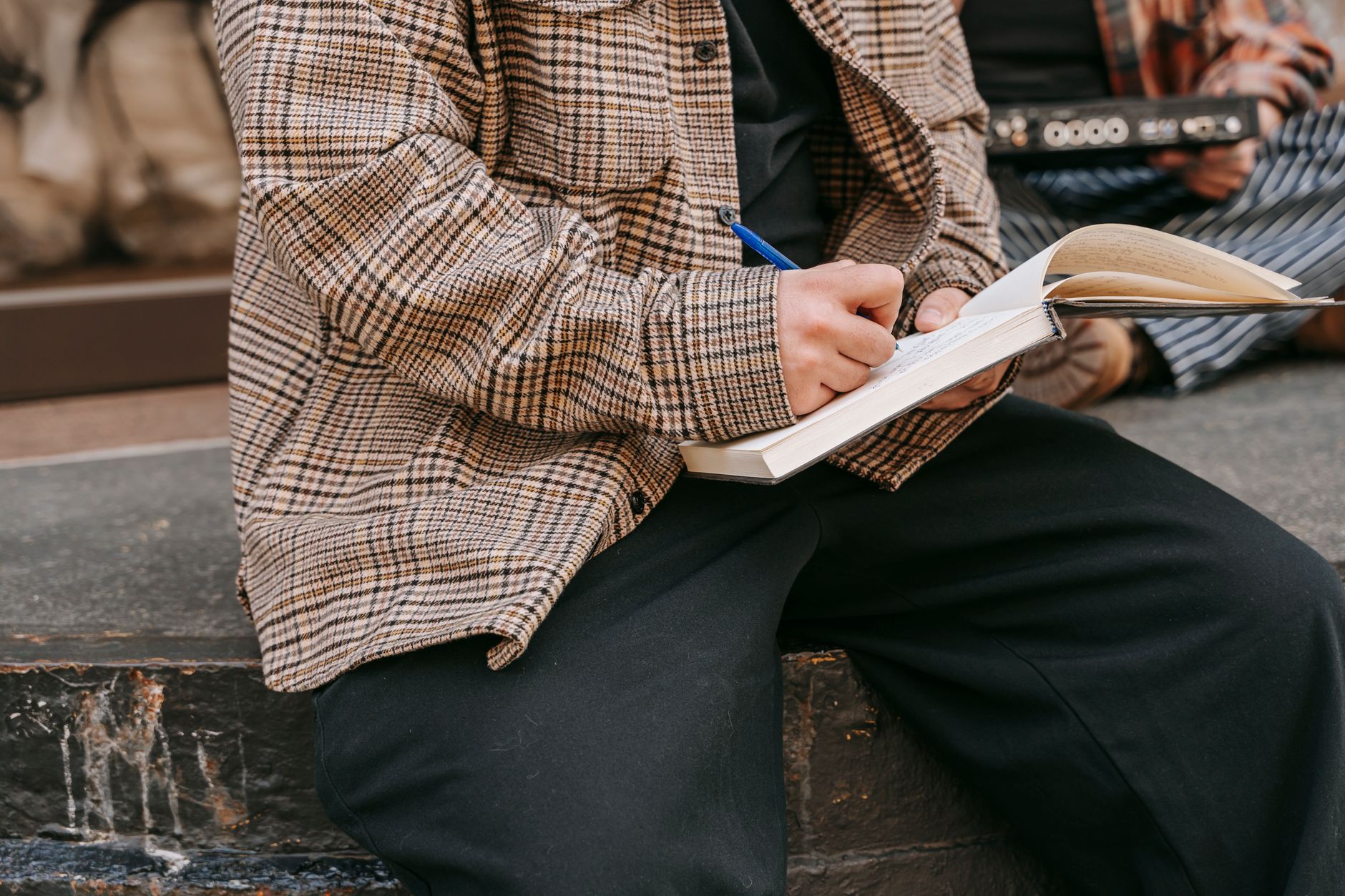 Ilustrasi: Close-up of a person writing in a notebook with a pen, sitting outdoors on a sunny day.