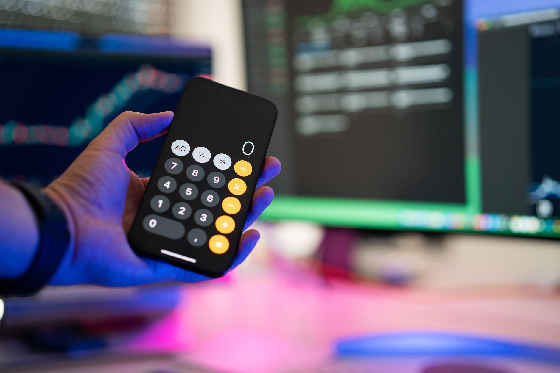 Ilustrasi: A person uses a smartphone calculator app in a modern office with computer screens displaying charts.
