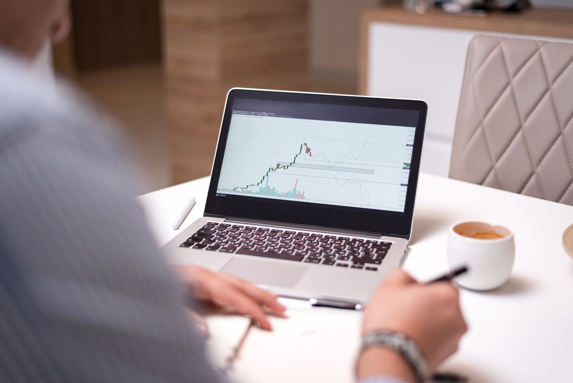 Ilustrasi: A person in a home office analyzing stock market data on a laptop with a coffee cup beside them.