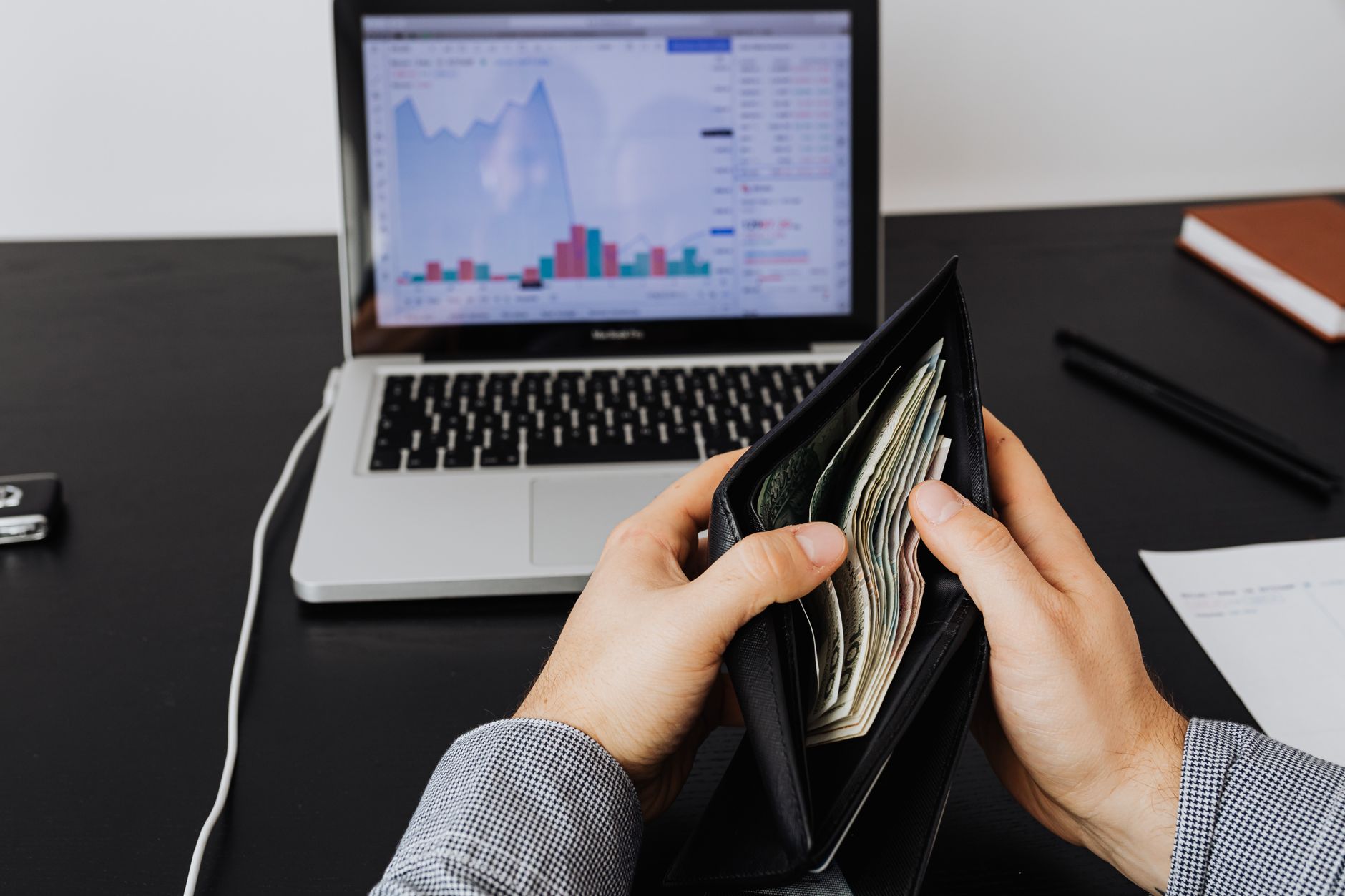 Ilustrasi: Man holding a leather wallet with bills in front of a laptop displaying financial graphs.