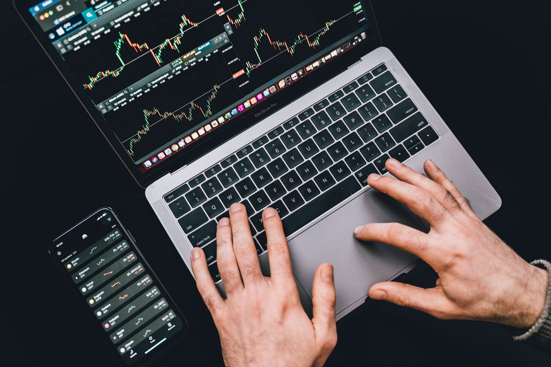 Ilustrasi: Hands typing on a laptop displaying stock market charts and a smartphone with financial apps.