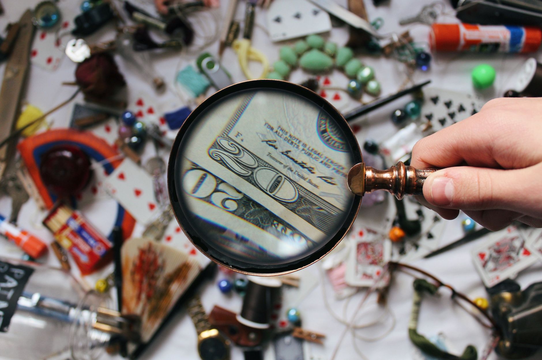 Ilustrasi: A hand holding a magnifying glass focusing on a twenty dollar bill amidst various scattered objects.