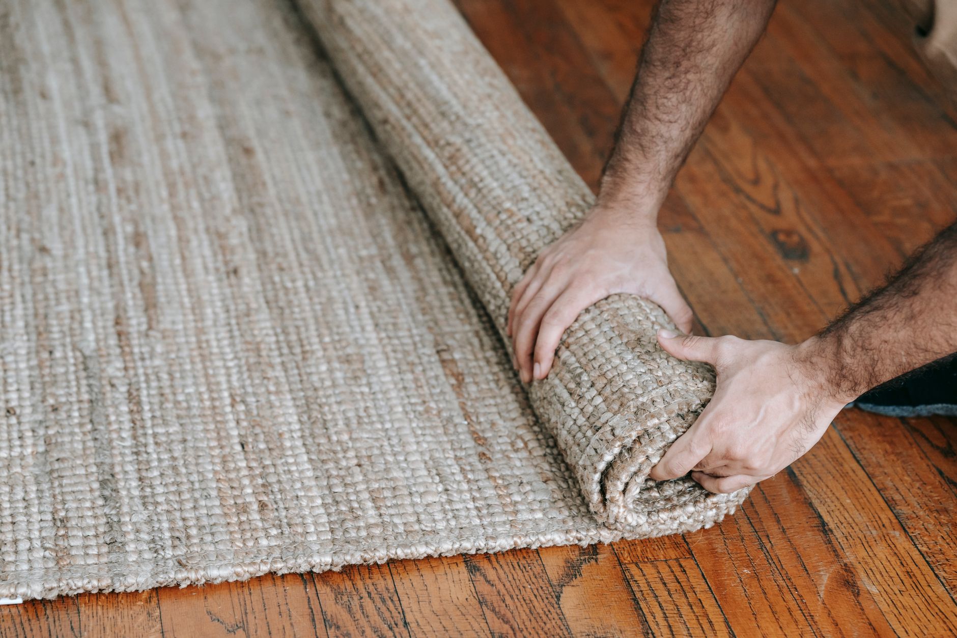 Ilustrasi: A man rolls up a textured woven rug on a wooden floor, indicating moving or cleaning.
