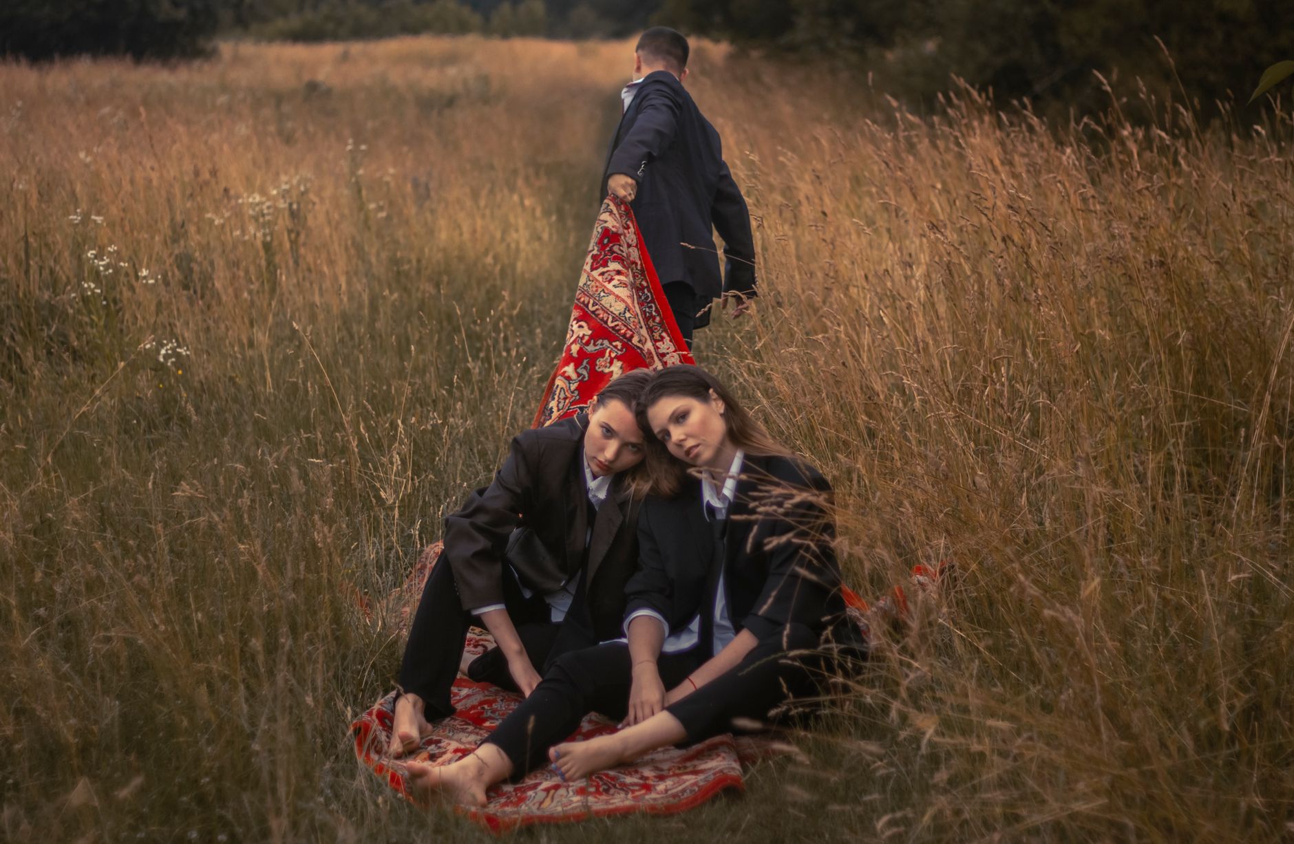 Ilustrasi: Two women sitting on a rug in a lapangan while a man pulls it, creating a conceptual outdoor scene.