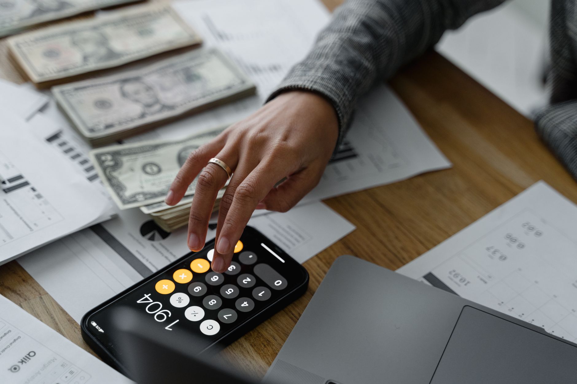 Ilustrasi: Close-up of a person using a smartphone calculator amid money and financial documents on a wooden table.