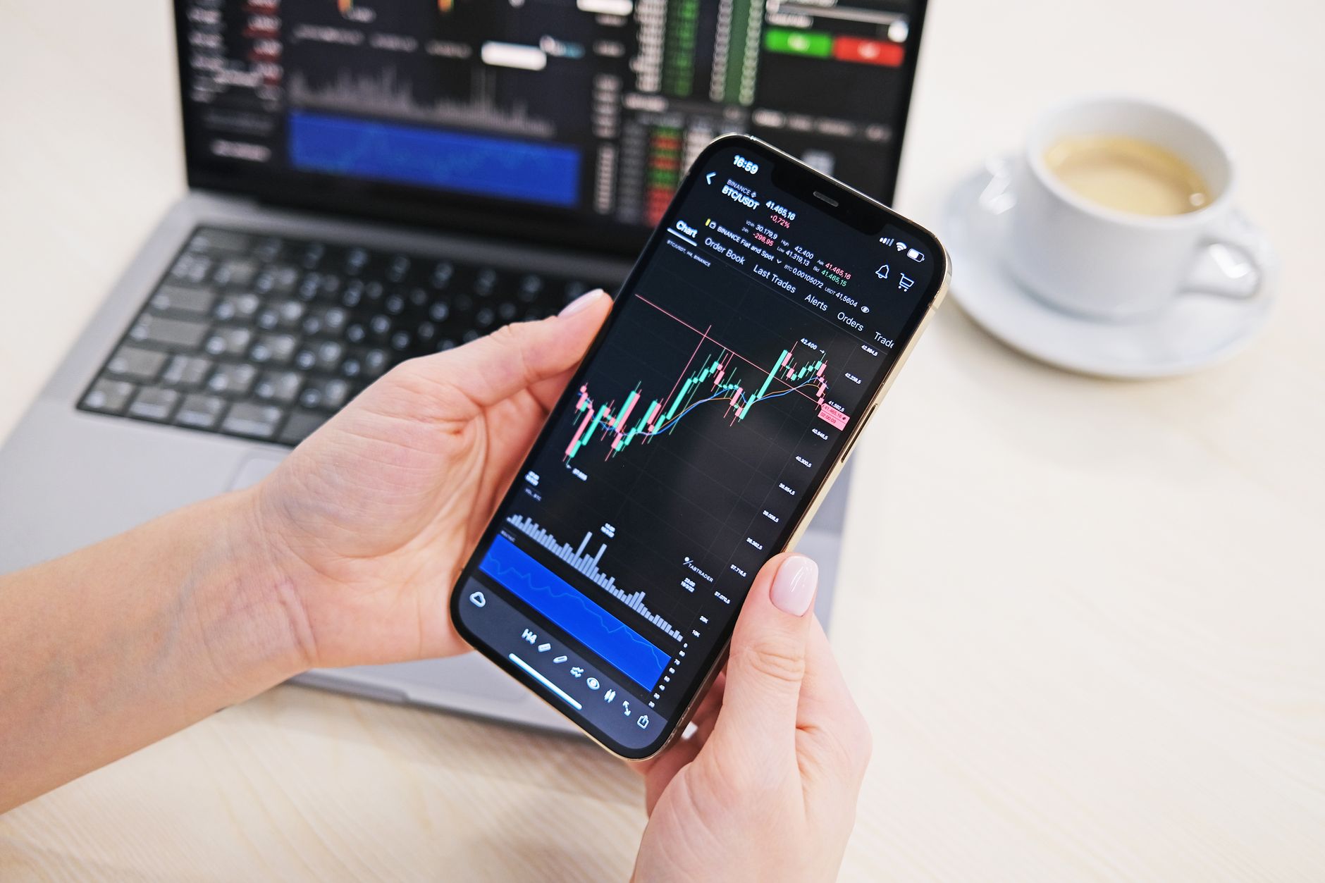 Ilustrasi: Close-up of hands holding smartphone displaying stock charts with laptop in the background on a desk.