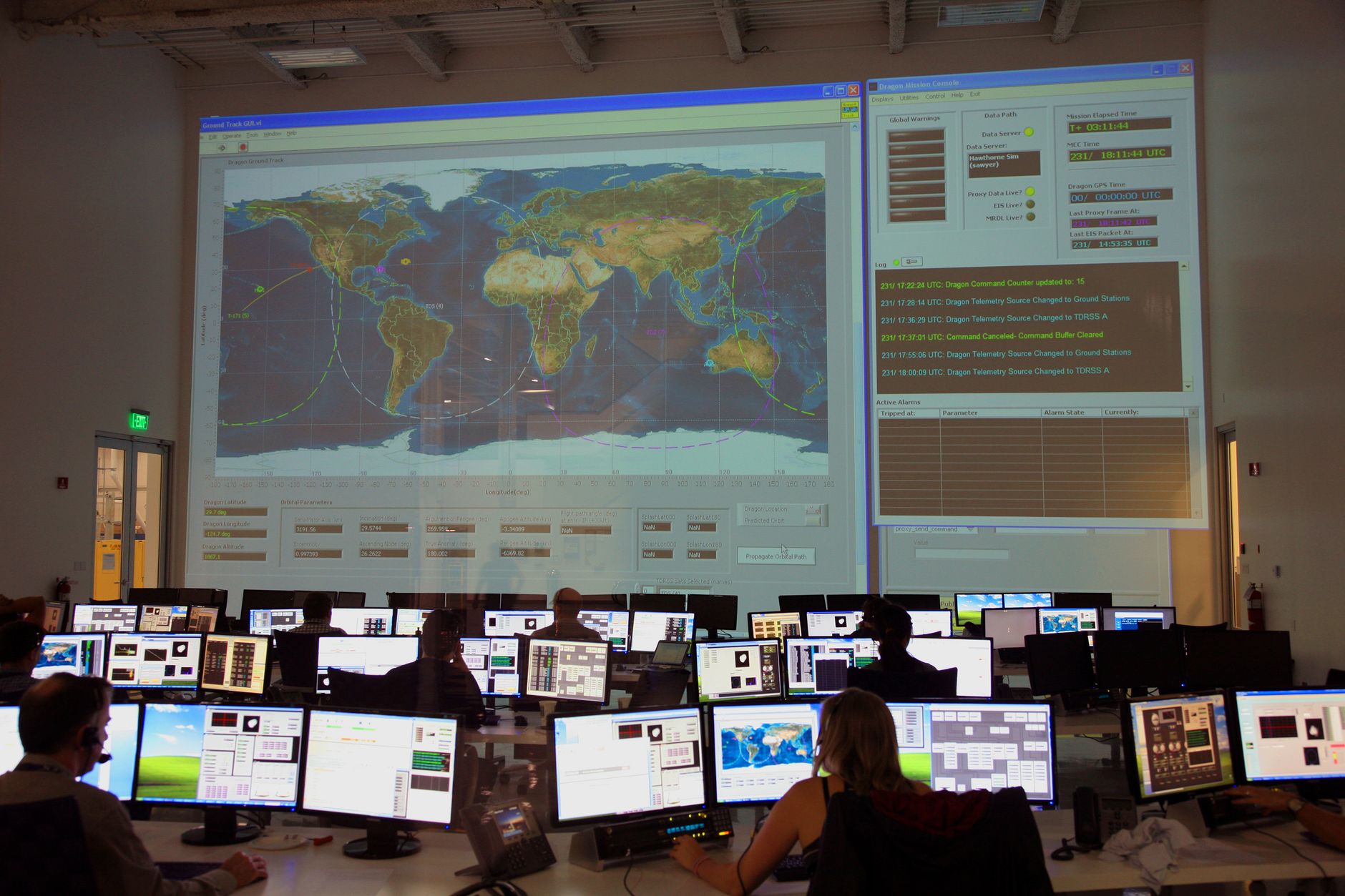 Ilustrasi: Back view of unrecognizable employees working on computers in flight control room with big interactive map on wall and analyzing data