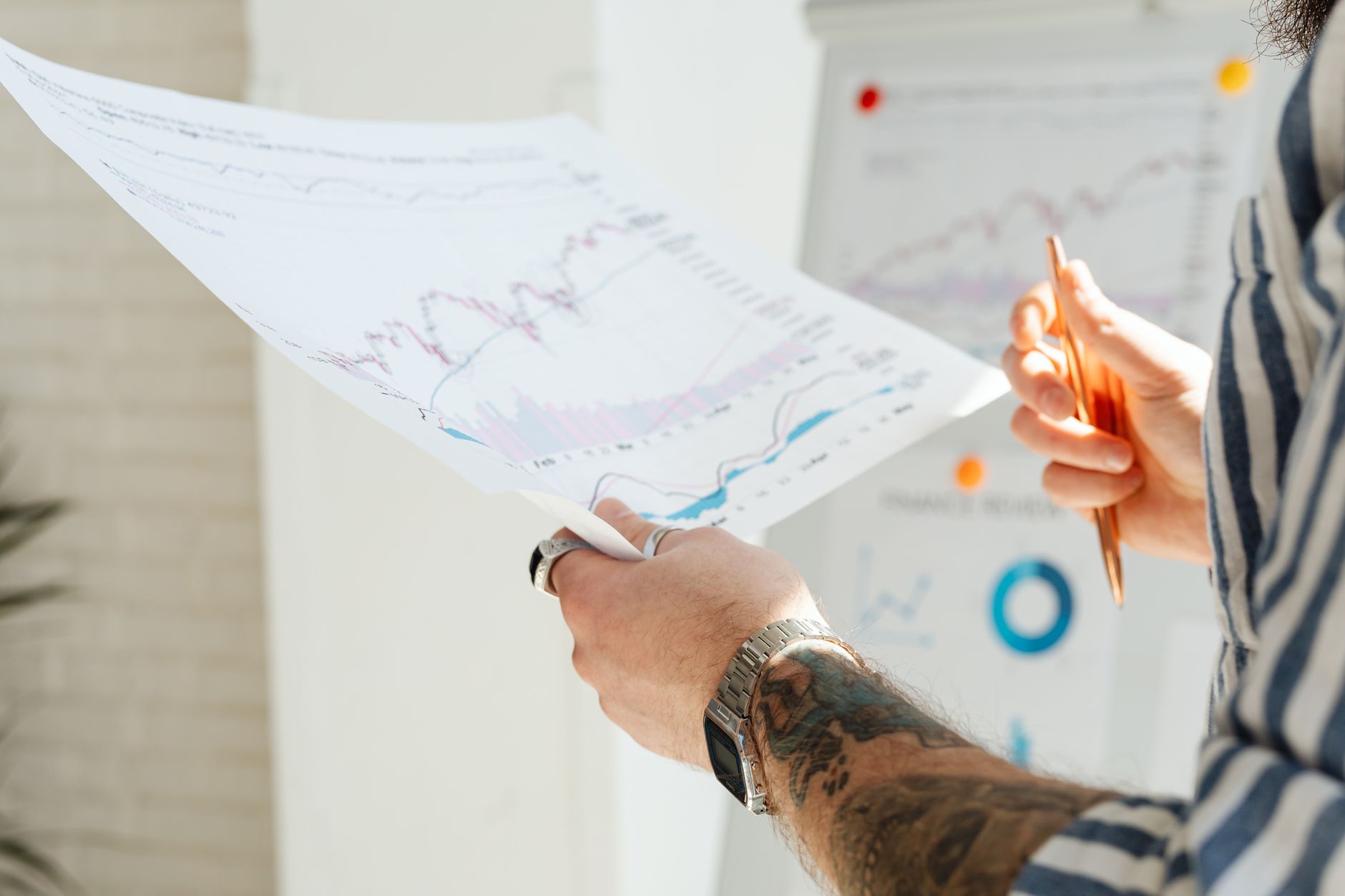 Ilustrasi: Close-up of a tattooed man's hands holding financial charts, with focus on data analysis and stock market trends.